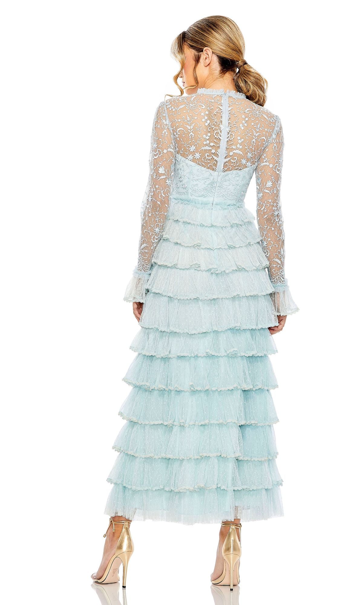 A woman stands with her back to the camera, wearing the Long Formal Dress 8003 by Mac Duggal in light blue lace with a sheer high neck, layered ruffles, and paired with metallic high-heeled shoes.